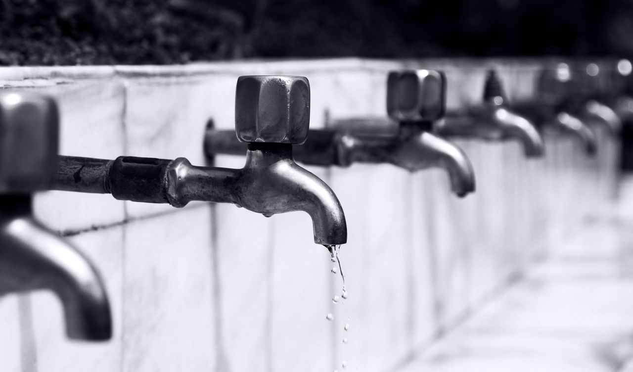 Black and white image of a row of faucets with water dripping, highlighting plumbing details.